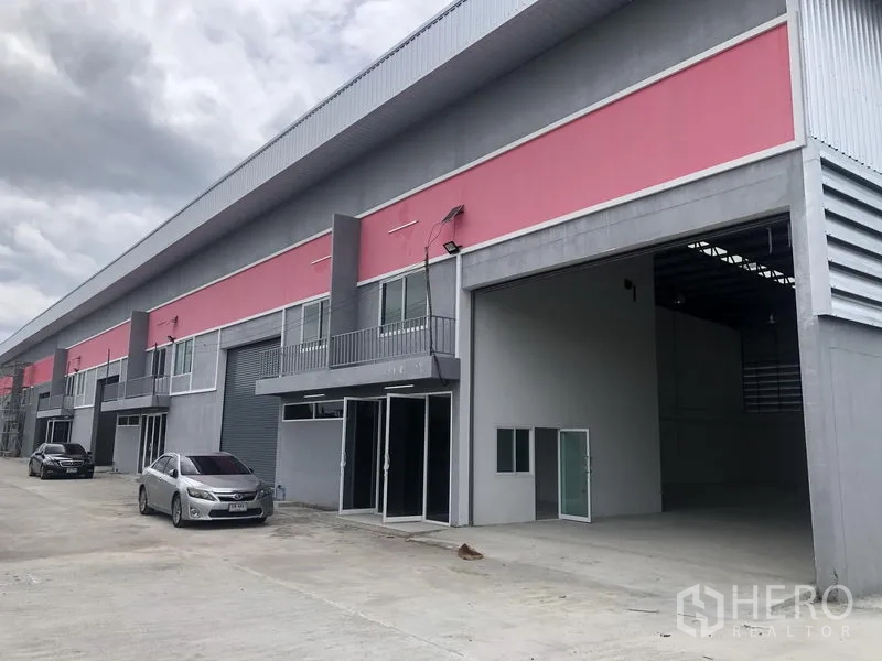 Factory for rent in Sai Noi, Nonthaburi - Row of factory-warehouse units with open loading bay and cars parked on a wide concrete driveway.