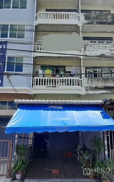 Retail Space for sale in Bang Na, Bangkok - Four-storey shophouse retail frontage with blue awning, roll-up shutter, balconies and potted plants in Bang Na.