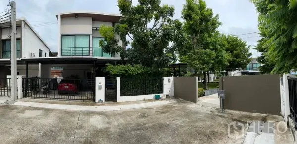 Detached House for sale in Samut Prakan - Wide street view of the gated property entrance and leafy surroundings.
