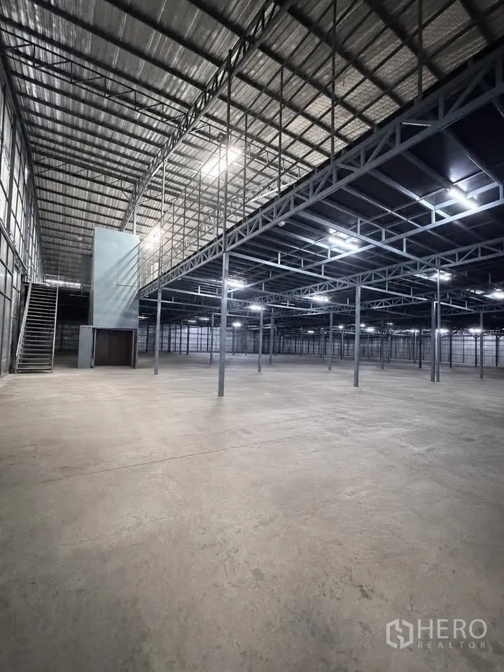 Warehouse for rent in Bang Khun Thian, Bangkok - Ground floor view with steel columns, mezzanine platform, stairs and bright overhead lights.