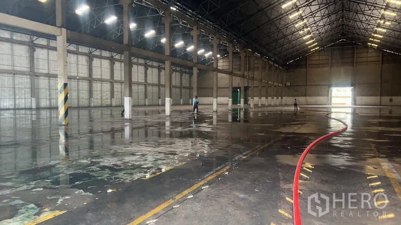 Warehouse for rent in Mueang Pathum Thani, Pathum Thani - Wide warehouse aisle with wet floor and a visible fire‑hose line near a loading door.