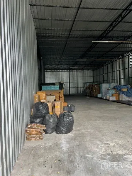 Warehouse for rent in Lat Phrao, Bangkok - High-ceiling metal-clad warehouse interior in Lat Phrao with concrete floor, fluorescent lighting, and stacks of boxes along the wall.