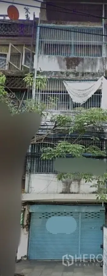 店屋 出租在Watthana, Bangkok - 多层商铺外立面，蓝色卷闸门，金属防护栏，风化混凝土立面，街边树木与电缆可见。