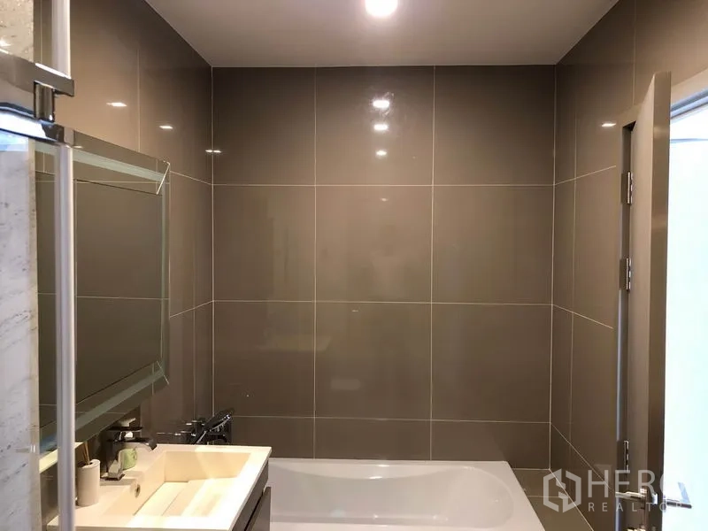 Condominium for rent in Watthana, Bangkok - Contemporary bathroom with bathtub, large beige wall tiles, vanity, and mirror.