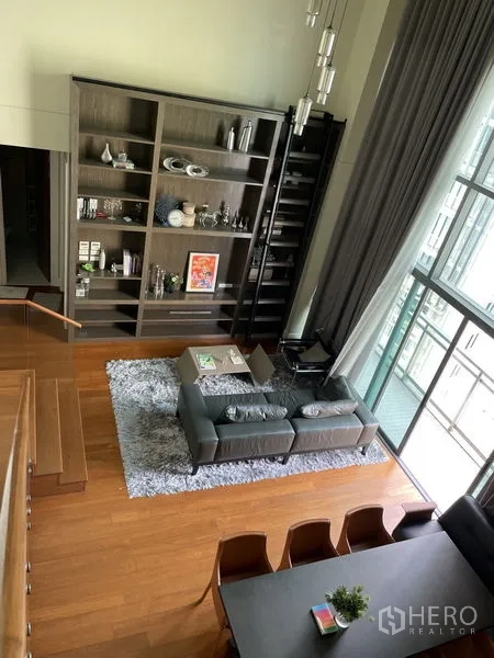 Condominium for rent in Khlong Toei, Bangkok - Open-plan lounge with tall built-in bookcase and ladder, pendant lights, gray sofa on a shag rug, and expansive glass windows.