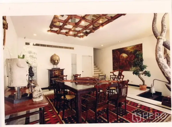 Townhouse for rent or sale in Watthana, Bangkok - Elegant dining room with carved wood ceiling feature, art-lined walls, and staircase beside an indoor water feature.