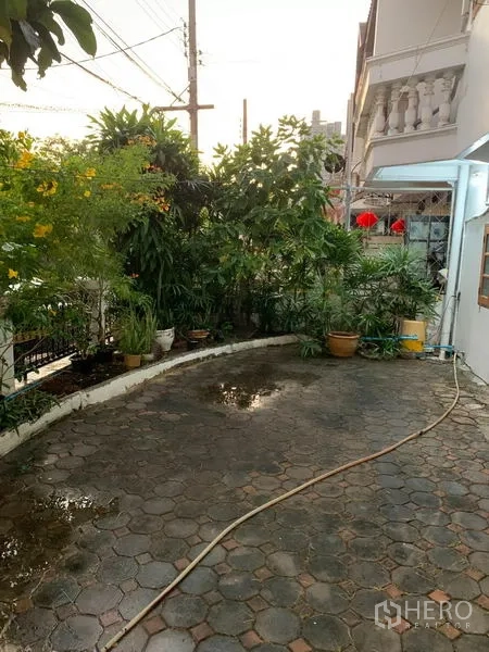 联排别墅 出租或出售在Phra Khanong, Bangkok - 前院弧形铺装地面，水管与繁茂热带绿植沿篱笆分布。