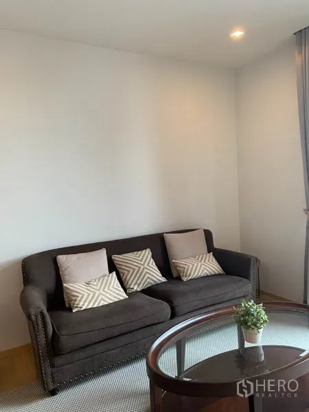 Condominium for rent in Phaya Thai, Bangkok - Cozy living room with a dark sofa, patterned cushions and a glass coffee table in a bright condo.