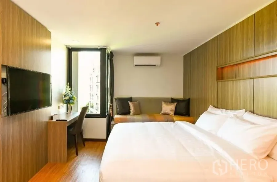 Shophouse for rent in Watthana, Bangkok - Bright modern room with wooden built-ins, wall-mounted TV, desk by a tall window, lounge seating, and air conditioner.