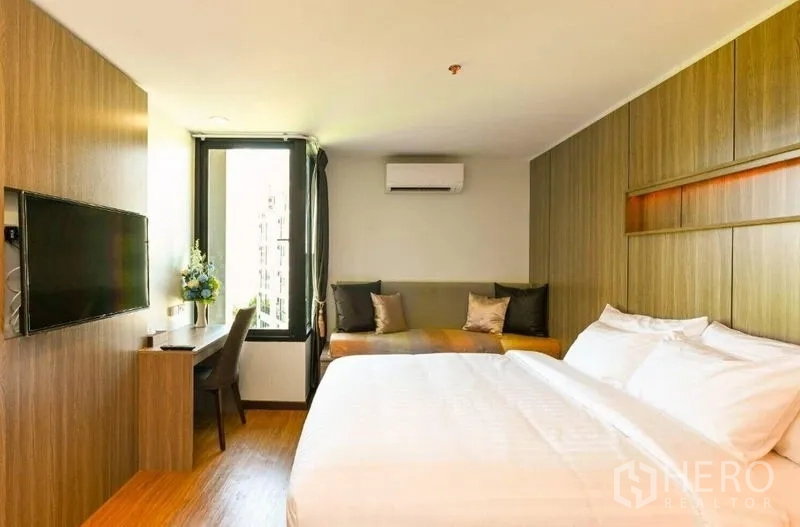 Shophouse for rent in Watthana, Bangkok - Bright modern room with wooden built-ins, wall-mounted TV, desk by a tall window, lounge seating, and air conditioner.