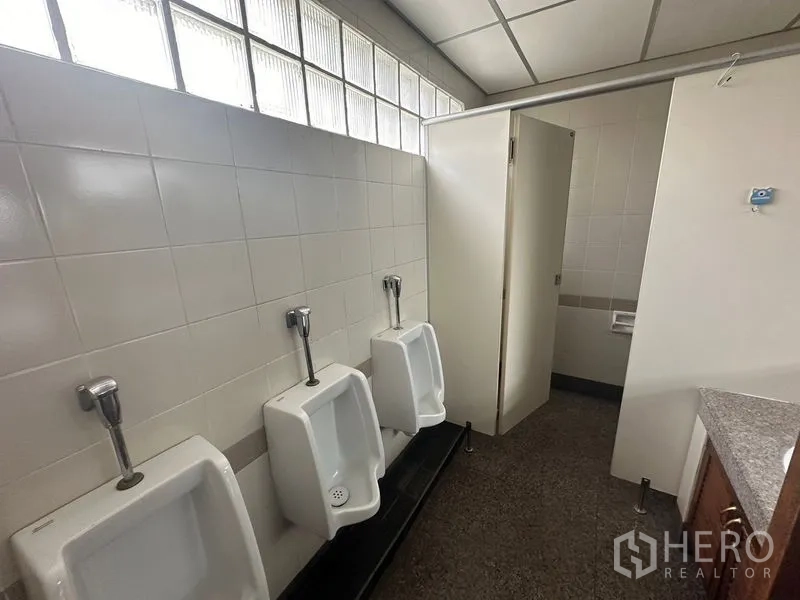 办公空间 出租 - 办公楼男厕，三个挂墙式小便斗，瓷砖墙面，隔间与玻璃砖采光窗。