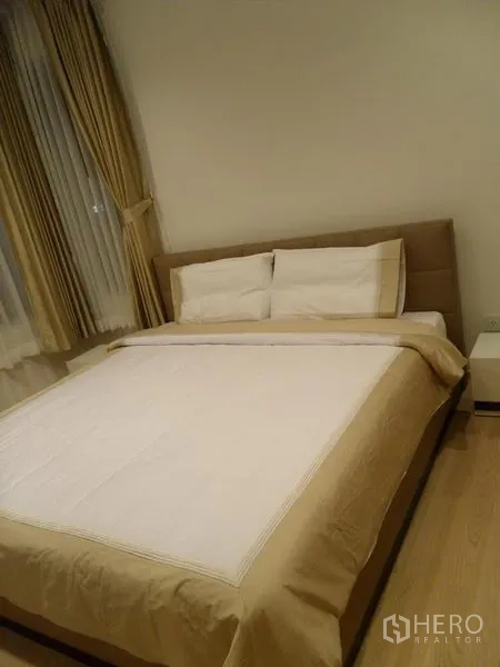 Condominium for rent in Khlong Toei, Bangkok - Cozy bedroom with double bed, beige bedding, bedside table and large window with drapes.