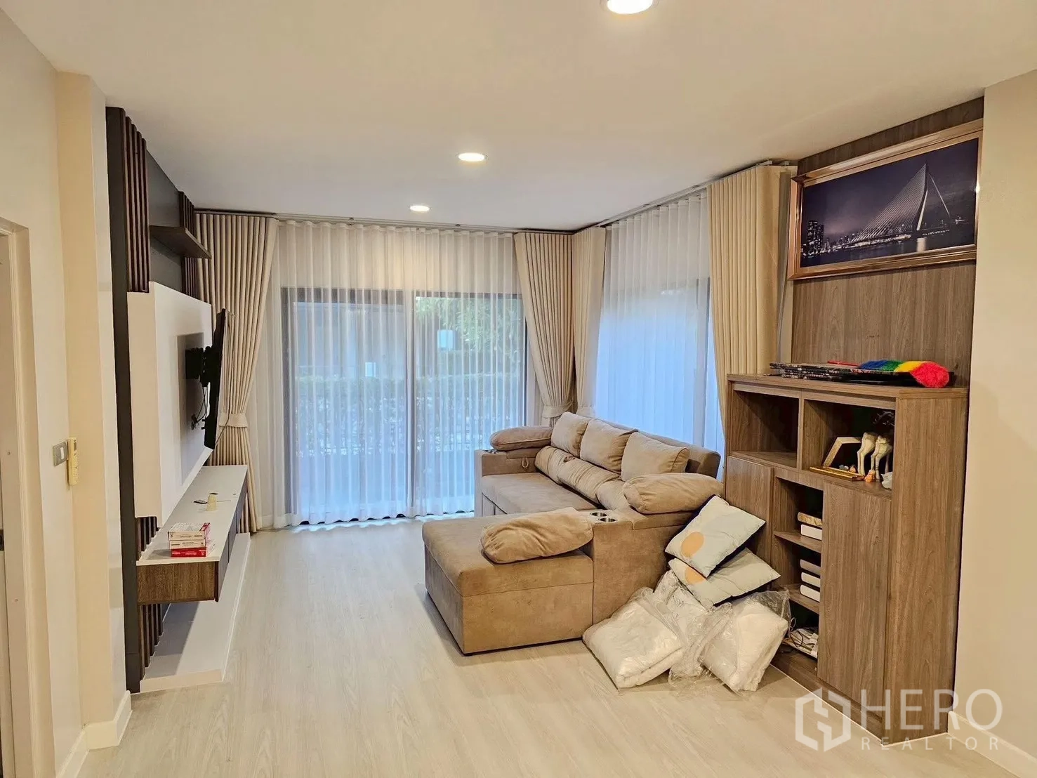 独立式住宅 出售在Bang Phli, Samut Prakan - 明亮客厅，L型沙发，电视墙与内置收纳柜，落地窗帘面向花园。