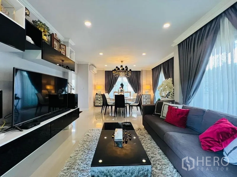 Detached House for rent or sale in Bang Khen, Bangkok - Open-plan living and dining room with chandelier, grey curtains, large TV and glossy tile floor.