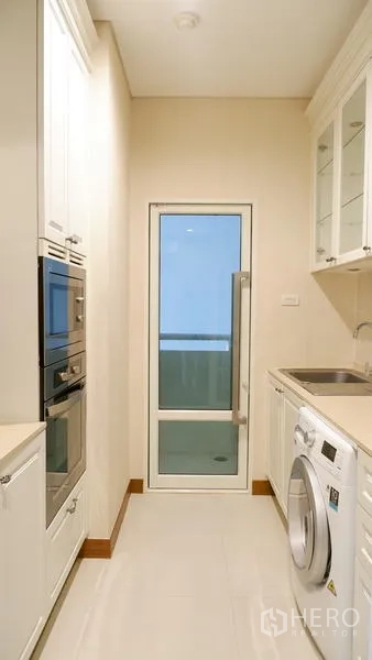 Condominium for rent in Watthana, Bangkok - Galley kitchen with white cabinetry, sink, and door to the utility balcony.