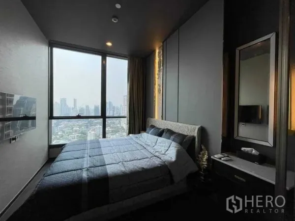 Condominium for rent in Khlong Toei, Bangkok - Cozy bedroom with a large window framing the Bangkok skyline, upholstered bed and wall‑mounted TV.