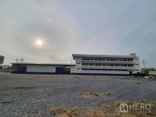 Factory for rent in Bang Bua Thong, Nonthaburi - Wide gravel yard facing a multi-storey factory and office block under a hazy sky.