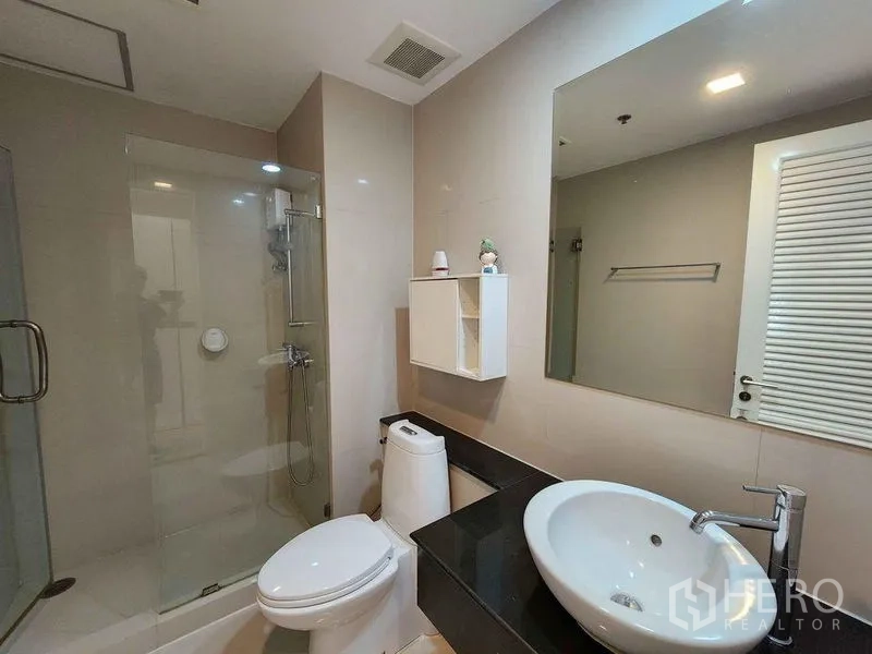 Condominium for rent in Khlong Toei, Bangkok - Modern bathroom with glass-enclosed shower, vanity sink and toilet in neutral tones.