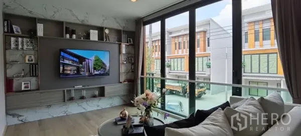 Townhouse for rent in Prawet, Bangkok - Second‑floor lounge with marble‑style wall, built‑in shelves, and wide sliding glass doors overlooking neighboring townhouses.