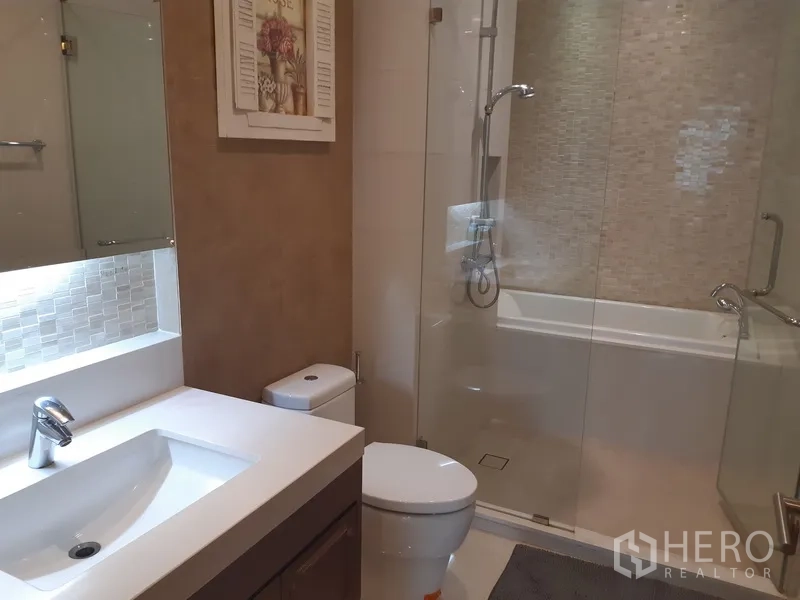 Condominium for rent in Khlong Toei, Bangkok - Modern bathroom with vanity, toilet, and a glass-enclosed shower over bathtub.