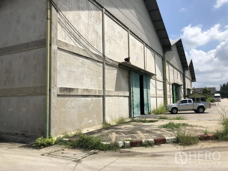 Warehouse for rent in Mueang Pathum Thani, Pathum Thani - Large concrete warehouse exterior with green sliding doors, loading apron and pickup parking in Mueang Pathum Thani.