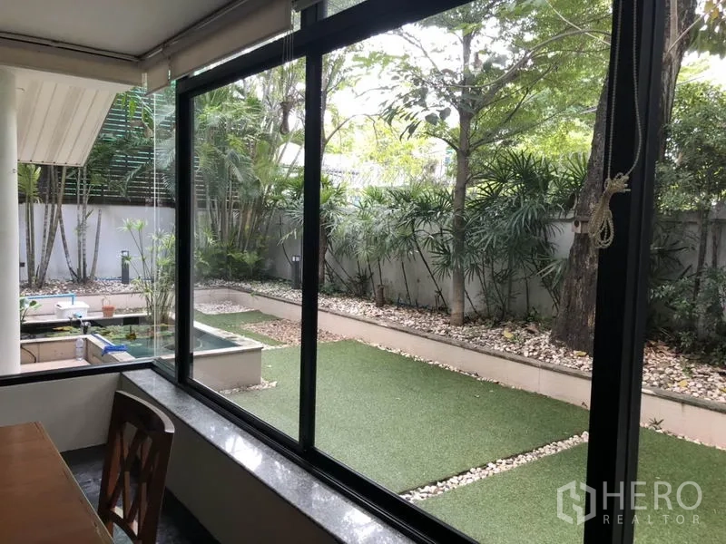 Detached House for rent in Bang Rak, Bangkok - Garden view through wide windows showing artificial lawn, raised planters, and trees.