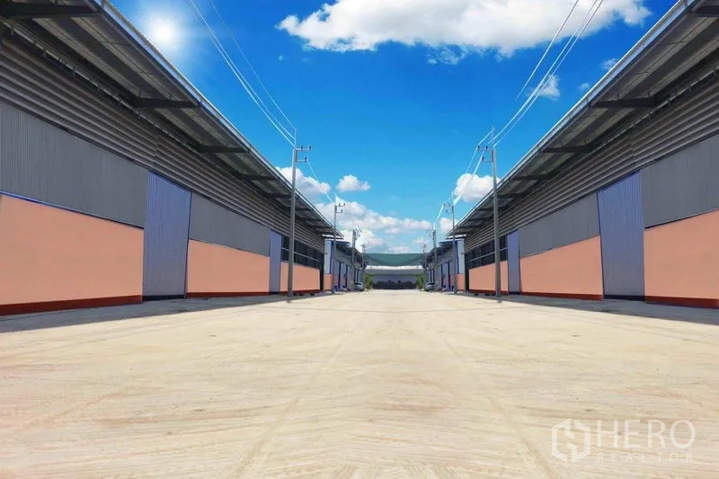 Warehouse for rent in Mueang Samut Sakhon, Samut Sakhon - Two rows of modern warehouse buildings facing a wide concrete driveway with power lines under a bright blue sky.