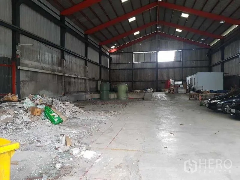 货栈 出租在Thung Khru, Bangkok - 仓库内部空间宽敞，红色钢梁挑高屋顶，混凝土地面，侧边有储物与水罐。
