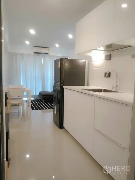 公寓 出租在Watthana, Bangkok - 开放式小厨房，白色橱柜与不锈钢水槽，大冰箱，前方连接餐区与客厅。