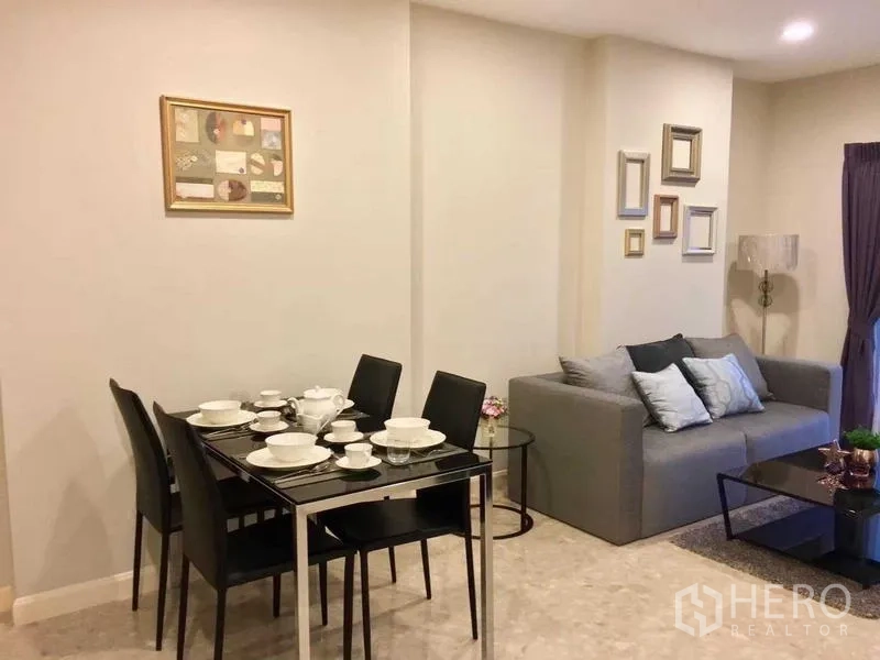 Condominium for rent in Khlong Toei, Bangkok - Dining and living space set with tableware and a gray sofa in warm tones.