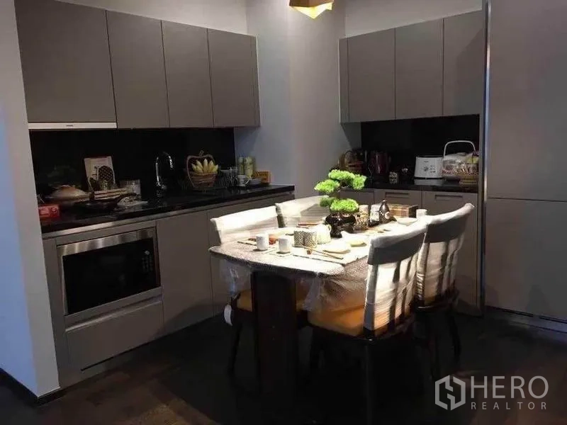 Condominium for rent in Watthana, Bangkok - Modern kitchen with matte gray cabinets, black stone countertop, and a four-seat dining table with bonsai centerpiece.