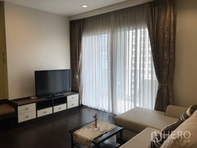 คอนโด ให้เช่าในRatchathewi, Bangkok - ห้องนั่งเล่นสว่าง โต๊ะวางทีวี พื้นไม้เข้ม และผ้าม่านโปร่งสูงจรดเพดานเปิดสู่ระเบียงวิวเมือง