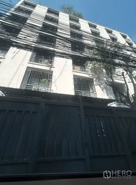 Warehouse for rent in Khlong Toei, Bangkok - Close view of a gated concrete facade with security grilles, tree shade, and dense power lines, showing a secure warehouse entrance in central Bangkok.
