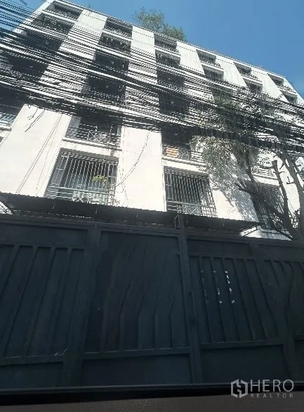 คลังสินค้า ให้เช่าในKhlong Toei, Bangkok - มุมมองด้านหน้าประตูโกดังคอนกรีตพร้อมเหล็กดัด ปกคลุมด้วยร่มเงาต้นไม้ และสายไฟหนาแน่น ใจกลางกรุงเทพฯ
