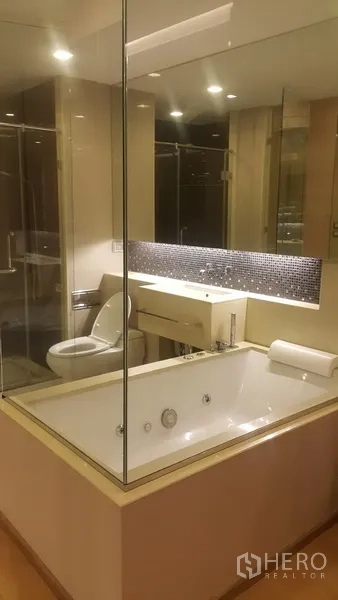 公寓 出租在Ratchathewi, Bangkok - 现代浴室，玻璃围合的浸泡浴缸、盥洗台和马桶，灯光柔和。