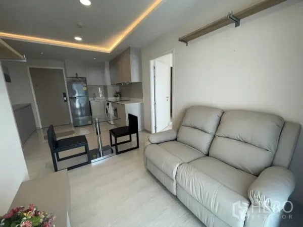 Condominium for rent in Khlong Toei, Bangkok - Bright living room with grey leather sofa, glass dining table, and open kitchen with fridge and washer.