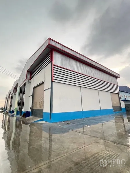 Warehouse for rent in Bang Phli, Samut Prakan - Corner view of the warehouse with roller shutters and a rain-wet concrete yard.