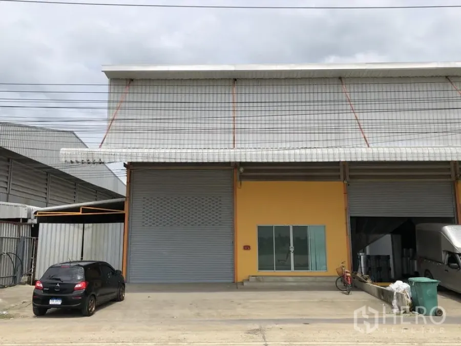 货栈 出租在Lat Lum Kaeo, Pathum Thani - 仓库正面，双卷帘门、玻璃办公室入口与宽阔车道停车区。