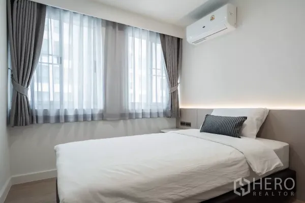 อพาร์ทเมนท์ ให้เช่า - ห้องนอนอบอุ่น เตียงขนาดมาตรฐาน หน้าต่างบานใหญ่พร้อมผ้าม่านสีเทา และเครื่องปรับอากาศติดผนัง