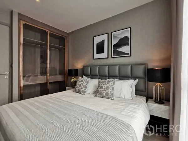 Condominium for sale in Watthana, Bangkok - Cozy bedroom with upholstered headboard, bedside lamps, framed art, and a sliding-door wardrobe in neutral tones.
