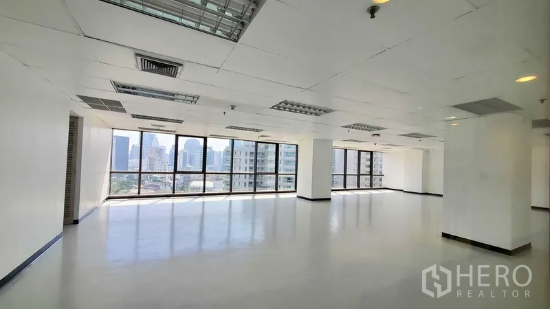办公空间 出租在Watthana, Bangkok - 明亮的开放式办公室，落地窗引入自然光与城市天际线，格栅吊顶配荧光灯和空调出风口。