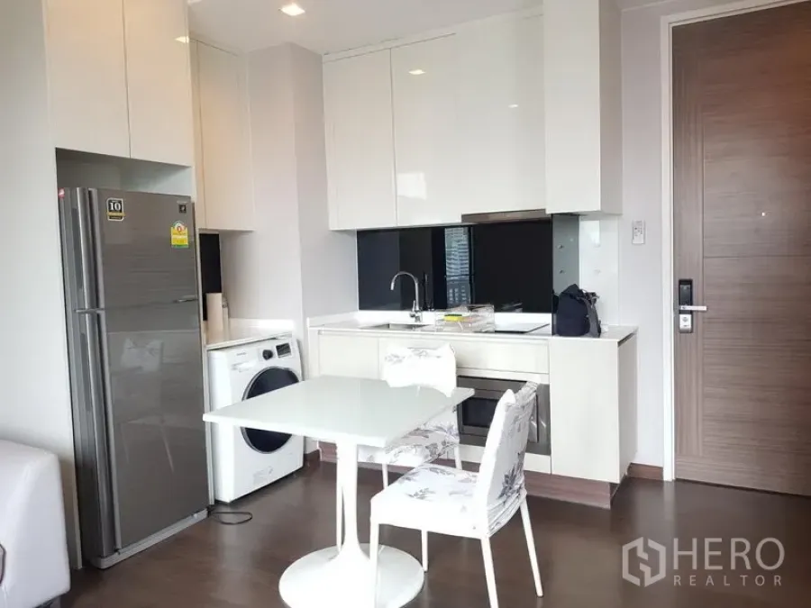 Condominium for rent in Ratchathewi, Bangkok - Modern kitchenette with glossy white cabinets, built-in oven, refrigerator and washing machine beside a small dining table.