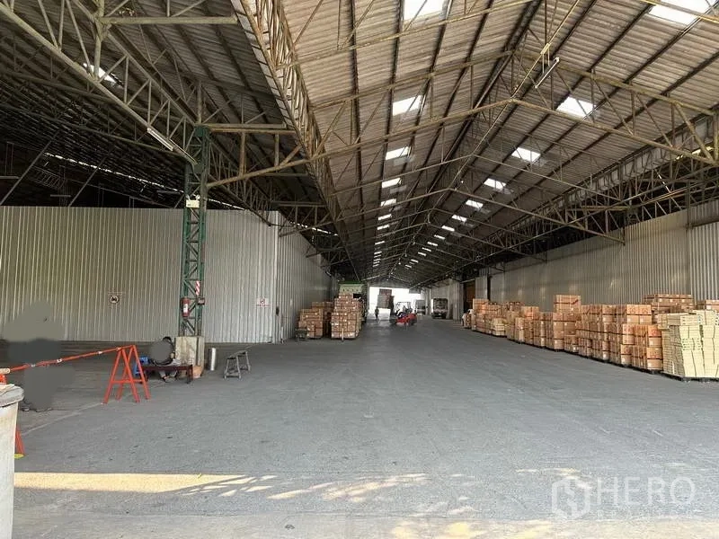 Warehouse for rent in Mueang Pathum Thani, Pathum Thani - Spacious warehouse interior with high steel truss roof, skylights, and pallet storage along both sides.