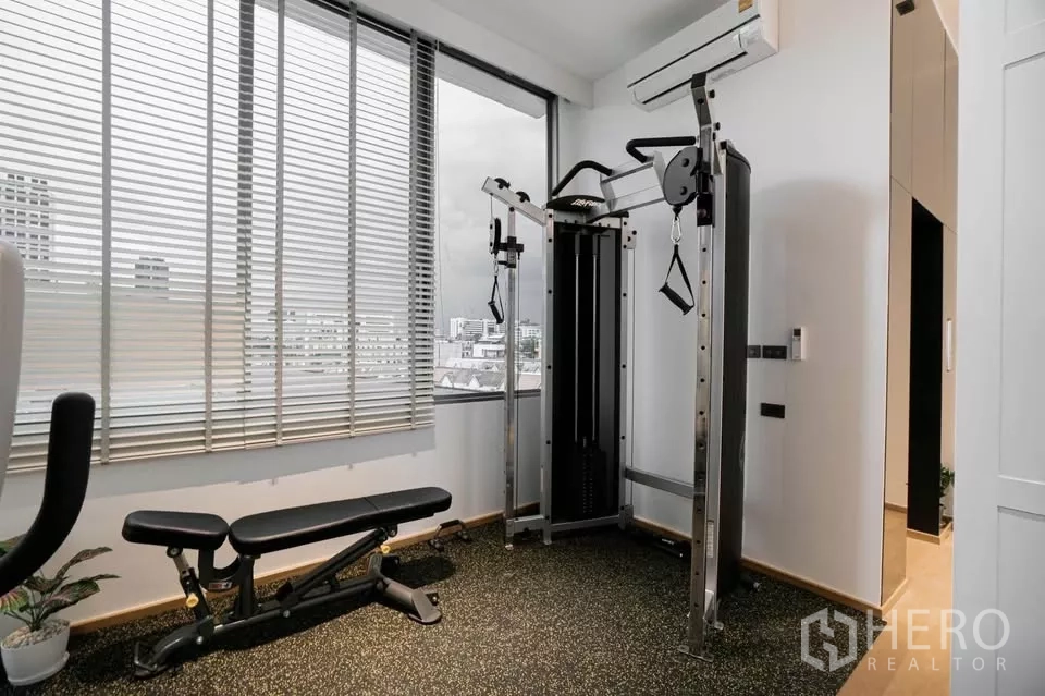 Townhouse for sale in Chatuchak, Bangkok - Home gym with bench, exercise machine and city views through blinds.