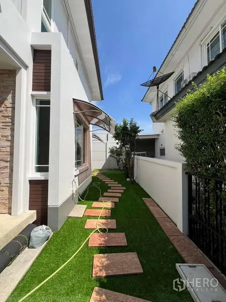独立式住宅 出租在Bang Kapi, Bangkok - 白色外墙旁的侧院草坪与踏步石小径并设遮雨棚。