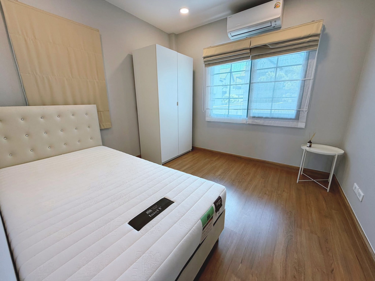 Detached House for rent in Bang Phli, Samut Prakan - Bright bedroom with white upholstered bed, wardrobe, wood floor, air conditioner and curtained window.