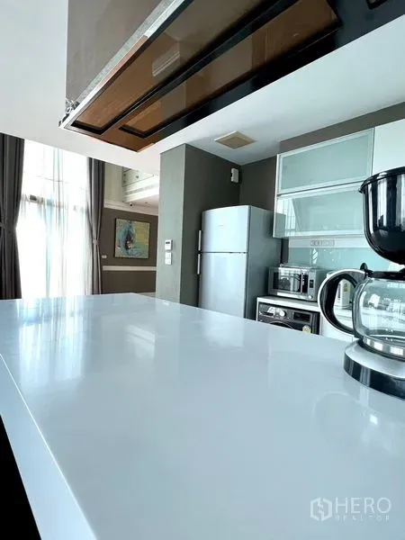 Condominium for rent in Khlong Toei, Bangkok - White breakfast bar and countertop in a bright kitchen with coffee maker and tall curtains.