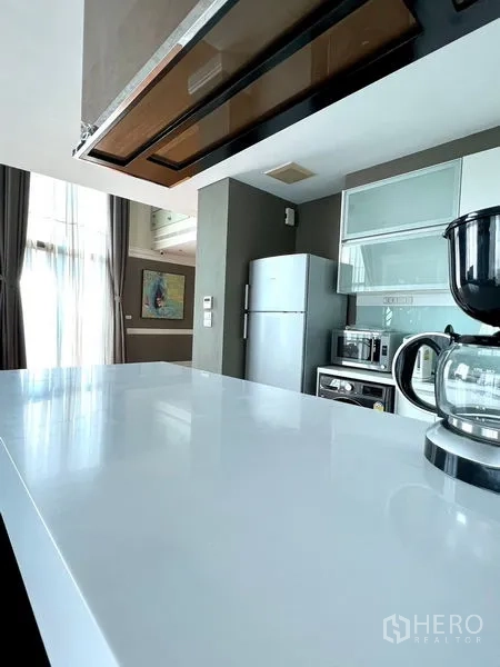 Condominium for rent in Khlong Toei, Bangkok - White breakfast bar and countertop in a bright kitchen with coffee maker and tall curtains.