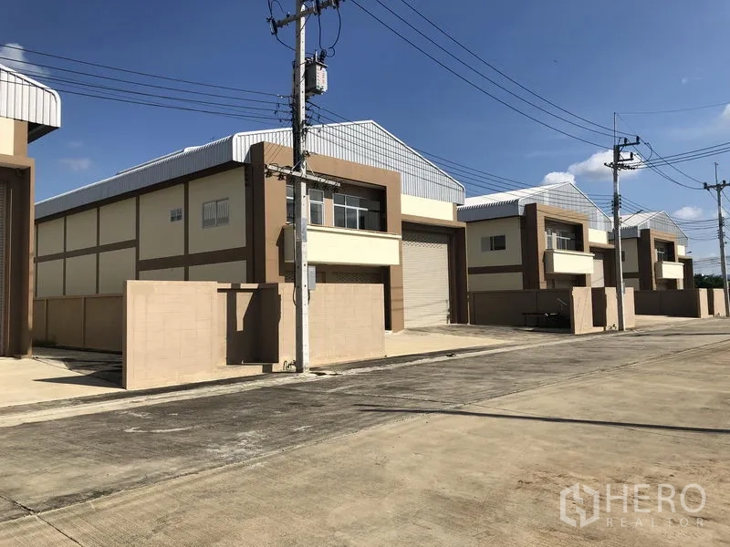 Factory for rent or sale in Pathum Thani - Row of similar warehouse units along a wide internal road with power poles and gated fronts.