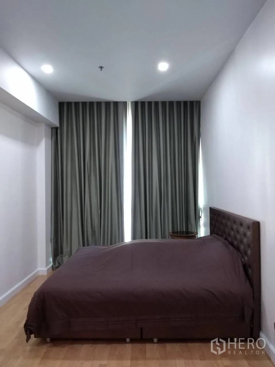 Condominium for rent in Khlong Toei, Bangkok - Primary bedroom with tufted headboard, dark blackout curtains, wood floors, and recessed lighting.